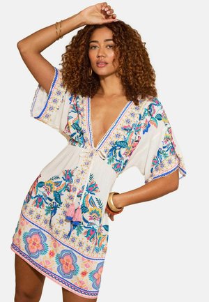 Kihara juustega naine, kes kannab valget lühikest kleiti lillemustris koos tutidega, poseerides ühe käe üles tõstetud ja teise käega puusal.