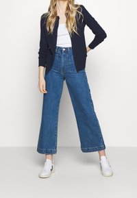 Modré džíny s širokými nohavicemi a vysokým pasem s texturovaným povrchem, doplněné bílým tílkem a tmavě modrým kardiganem na knoflíky.