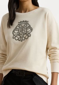 Felpa beige dal taglio ampio, con dettagli di ricamo in argento intrecciati a spirale sul petto. Maniche lunghe e colletto a coste.