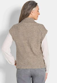 Un gilet sans manches beige léger et côtelé avec un col haut, porté sur une chemise blanche à manches longues. La vue de dos met en avant la douceur du tissu et l'ajustement.