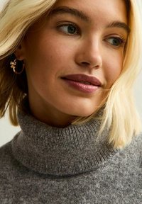 Grå stickad turtleneck-tröja med ett mönster, i kombination med guldörhängen i hoops med bladdekorationer, framhävt mot en ljus bakgrund.