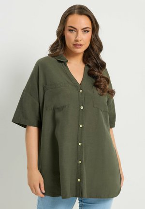 Vrouw met lang golvend haar, gekleed in een oversized, olijfgroen overhemd met korte mouwen en lichtblauwe spijkerbroek, staand tegen een effen achtergrond.