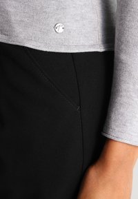 Top in maglia grigio chiaro con dettaglio logo argento, abbinato a pantaloni in tessuto nero con cuciture visibili e texture liscia.