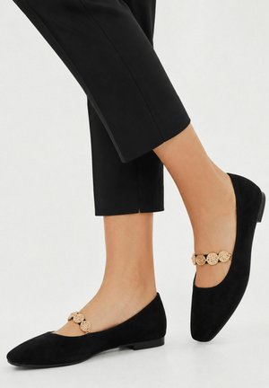 Zwarte platte schoenen met gouden sierband gedragen bij cropped zwarte broeken, waarbij voeten en onderbenen zichtbaar zijn tegen een effen achtergrond.