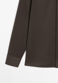 Donkerbruine langemouwshirt met een soepele textuur, knoopmanchetten en een iets ontspannen pasvorm. Stiksel details zijn zichtbaar langs de voorkant.