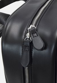 Borsa da viaggio in pelle nera con una zip argento prominente, texture morbida e dettagli cuciti vicino ai tiranti della zip.