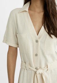 Kobieta ubrana w lnianą sukienkę w kolorze beżowym z krótkim rękawem, kołnierzykiem, zapięciem na guziki z przodu, kieszeniami na piersi i przewiązanym paskiem w talii, skróconą na ramionach i w talii.
