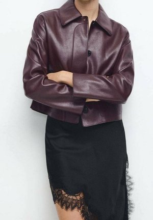 Personne portant une veste en cuir courte violet foncé avec des boutons noirs sur une robe nuisette noire bordée de dentelle, bras croisés devant un fond uni.