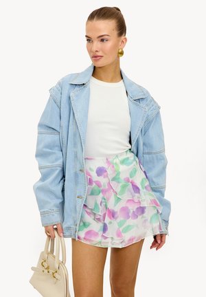 Femme portant une veste en denim bleu clair oversize, un haut blanc, une mini-jupe à volants floraux, des boucles d'oreilles dorées, tenant un sac à main beige, regardant sur le côté.
