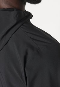 Chaqueta negra con una textura suave y mate. Presenta capucha, costuras de hombros estructuradas y un diseño limpio con un mínimo de detalles.