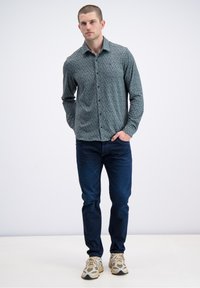 Overhemd met knopen in donkerblauwgroen met een bladmotief, gecombineerd met donkerblauwe spijkerbroek en lichtgekleurde sneakers, staand op een neutrale achtergrond.