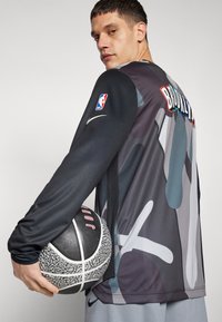 Sort og grå langærmet basketballtrøje med abstrakte mønstre. Har NBA-logo, teksten "BROOKLYN" og en basketball med tekstureret overflade.