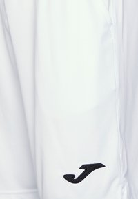 Białe spodenki sportowe z czarnym haftowanym logo "J" w pobliżu dolnego brzegu po prawej stronie.