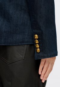 Mano appoggiata accanto alla tasca dei pantaloni in pelle nera, indossando una giacca di denim scuro con quattro bottoni dorati sul polsino.