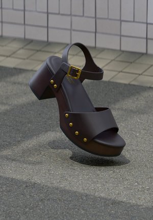 Sandale compensée en cuir marron foncé avec bride à la cheville, boucle dorée et clous dorés, posée sur un pavé texturé près d'un mur carrelé.