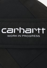 Czarna torba z tkaniny z białym logo "carhartt WORK IN PROGRESS", z widocznymi szwami i detalami uchwytu zapewniającymi pewne noszenie.