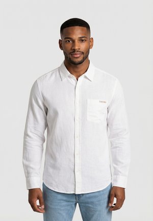 Uomo che indossa una camicia bianca a maniche lunghe con bottoni e taschino sul petto, e jeans azzurri, in piedi davanti a uno sfondo semplice.