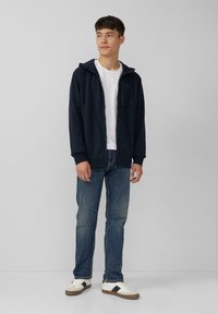 Jonge man in een marineblauwe ritssluiting hoodie, wit t-shirt, blauwe jeans en witte sneakers met zwarte strepen, staand tegen een effen achtergrond.