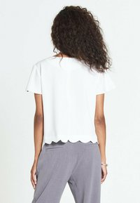 Top bianco corto con maniche corte, orlo scallop e tessuto dalla superficie liscia. Indossato con pantaloni grigi, con un design a schiena scoperta.
