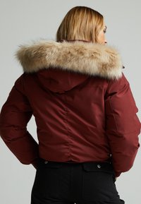 Burgundska jakna s lažnom krznom na kapuljači, koja ima glatku teksturu, kroj koji ističe figuru i izdržljive šavove duž leđa.