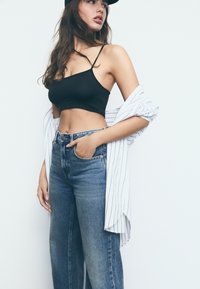 Svart kortad tanktop som matchas med höga blå jeans, med en randig öppen skjorta som hänger över ena armen.