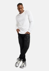 Hvid strikket sweater, sorte jeans og mønstrede høje sneakers. Sweateren har ribbede manchetter og kant.