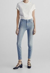 Lichtblauwe high-waisted jeans met een slim fit, gecombineerd met een witte t-shirt met opgerolde mouwen en zwarte sleehakken. Eenvoudige, casual uitstraling.