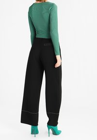 Green long-sleeve top with textured fabric, paired with high-waisted black wide-leg pants featuring white stitching and teal high heels.