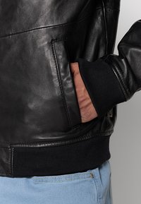 Giacca bomber in pelle nera con una tasca laterale, polsini e vita a coste. Texture liscia e design aderente. Leggera lucentezza.