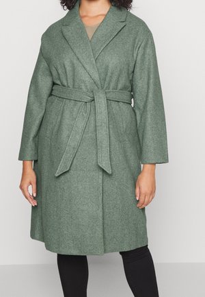 Grüner knielanger Wollmantel mit Gürtel und breiter Revers, getragen über einer schwarzen Hose an einer stehenden Person.