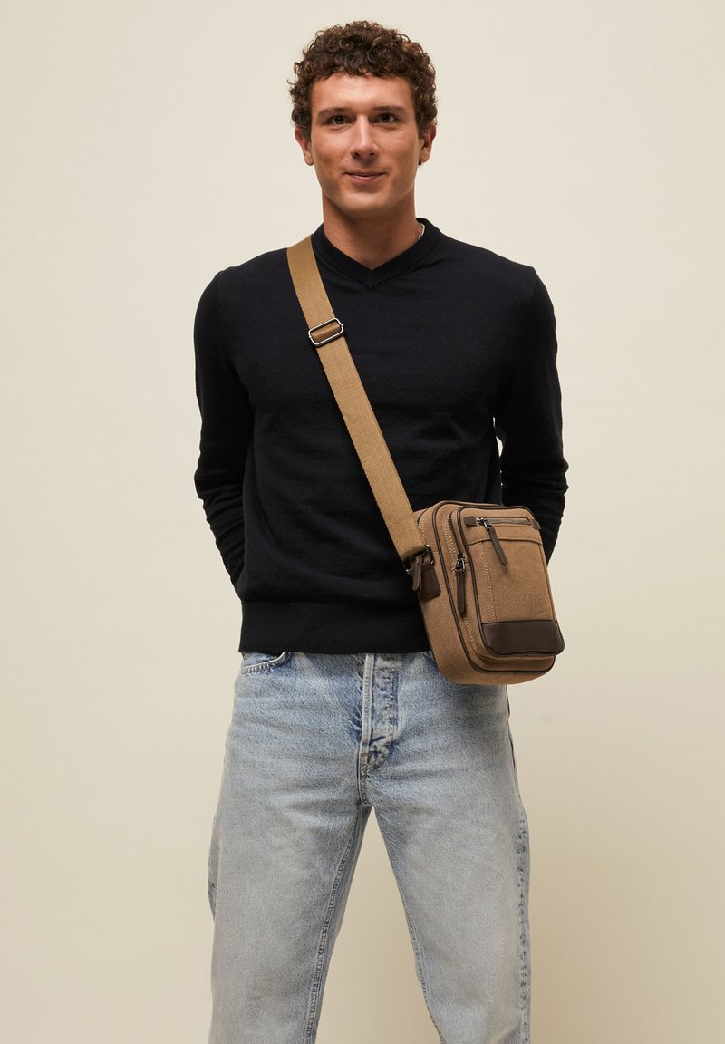 Jonge man met krullend haar die een zwarte trui, lichtblauwe spijkerbroek en een bruine crossbodytas draagt tegen een effen beige achtergrond.