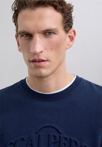 Sweatshirt bleu marine avec logo textuel en relief, col rond et tissu texturé. Le modèle a les cheveux courts et ondulés et fait face à l'avant.