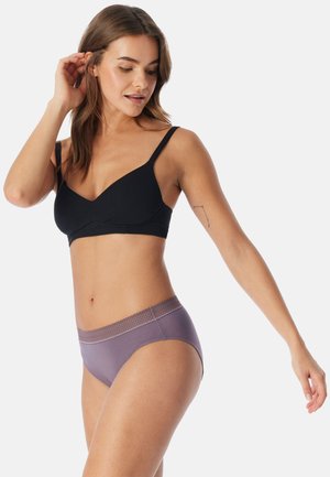 Frau trägt schwarzen BH und lila High-Waist-Unterwäsche, blickt nach unten, eine Hand nahe am Ohr, die andere seitlich ausgestreckt.