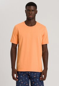 T-shirt a maniche corte in cotone arancione pastello, con scollo rotondo. Indossato con pantaloni corti blu navy a fantasia che mostrano disegni circolari.