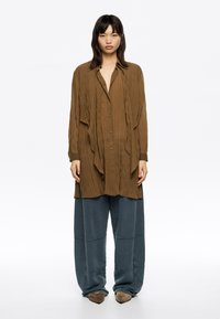Brown, textured long-sleeve shirt with button front, side slits, paired with loose gray-blue sweatpants and pointed brown shoes.