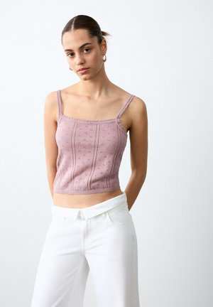 Mlada ženska z lasmi v figi, ki nosi obroče za ušesa, vijoličast pleteni top brez rokavov in bele hlače z visokim pasom, stoji pred enobarvnim ozadjem.