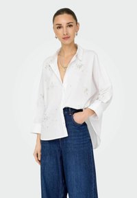 Camicia bianca con bottoni e decorazioni di perline, con colletto, maniche lunghe e vestibilità rilassata, abbinata a jeans a gamba larga blu scuro.