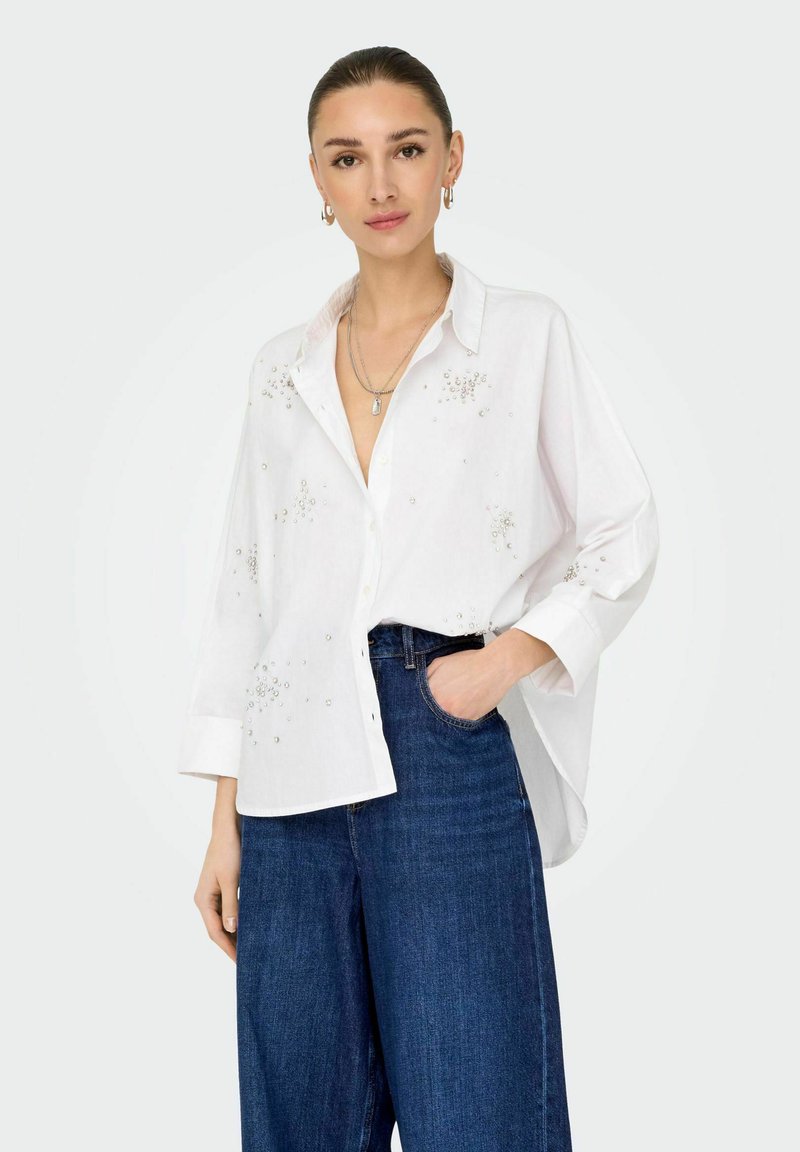 Camicia bianca con bottoni e decorazioni di perline, con colletto, maniche lunghe e vestibilità rilassata, abbinata a jeans a gamba larga blu scuro.