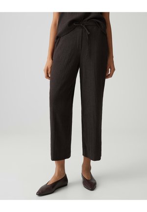 Donna che indossa pantaloni neri corti con trama e vita con coulisse, abbinati a scarpe basse scure con punta, su sfondo chiaro.