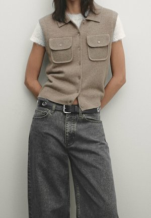 Osoba ubrana w beżową kamizelkę na guziki z klapkowymi kieszeniami, na białym, krótkim, puszystym swetrze oraz czarne, wysokowciskane, szerokie dżinsy z czarnym paskiem.