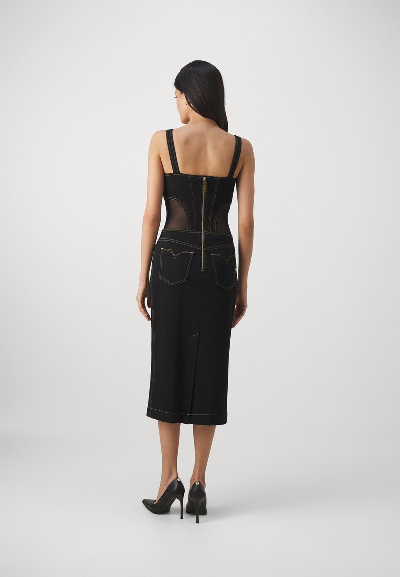 Femme portant une robe midi noire ajustée avec des panneaux latéraux transparents, des coutures dorées, une fermeture éclair au dos et des chaussures à talons noirs sur un fond blanc.