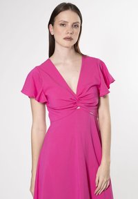 Fuchsia kjole med korte flæseærmer, en knyttet frontdetalje og en dyb V-udskæring. Lavet af letvægtsstof med glat tekstur.