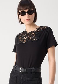 Sort T-shirt med intrikate sorte blonder detaljer ved udskæringen. Kombineret med højtaljede sorte jeans og et sort bælte med en sølvspænde.