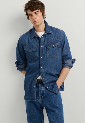 Junger Mann, der ein blaues Jeanshemd mit hochgekrempelten Ärmeln über einem weißen T-Shirt und blauen Jeans trägt, steht mit einer Hand in der Tasche vor einer schlichten Wand.