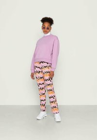 Sweatshirt roxo com gola redonda; calças estampadas em rosa, laranja e preto; ténis brancos; óculos de sol redondos; detalhes em tecido texturizado.