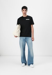 Juodas medvilnės marškinėliai su balta "Calvin Klein" logotipu, derinami su šviesiai mėlynais plačiais džinsais ir laikant balta tekstūruota rankinę.