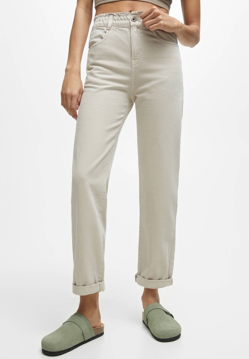 Femme portant un pantalon taille haute beige clair avec des revers retroussés et des chaussures vertes sans lacets, debout devant un fond blanc uni.
