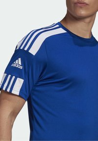 Niebieska koszulka sportowa z okrągłym dekoltem, wykonana z gładkiego materiału. Posiada białe poziome paski na ramionach oraz logo Adidasa na rękawie.