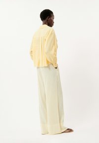 Chemise jaune à boutons avec un ourlet effiloché associée à un pantalon large à rayures verticales claires, vue de côté sur un fond uni.