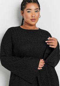 Mustapintainen yläosa pyöreällä kaula-aukolla, pitkät kellotaskut ja hienovarainen rypytetty kuvio. Kangas näyttää kevyeltä ja laskeutuu pehmeästi.